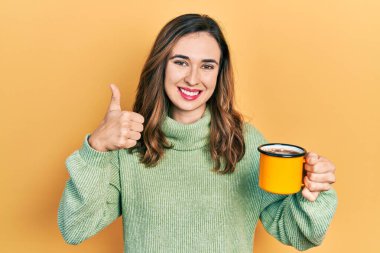 Genç İspanyol kız bir fincan kahve içiyor mutlu ve pozitif gülümsüyor, başparmağıyla mükemmel ve onay işareti yapıyor. 