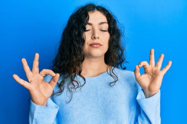 Kıvırcık saçlı, rahat giyinen, gözleri kapalı gülümseyen ve parmaklarıyla meditasyon yapan genç esmer kadın. yoga konsepti. 