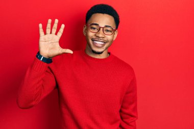 Genç Afro-Amerikalı günlük kıyafetler ve gözlük takmış beş numaralı parmaklarıyla işaret ederken kendinden emin ve mutlu bir şekilde gülümsüyor.. 
