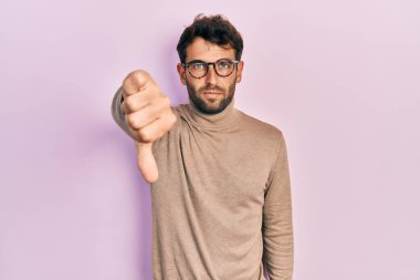 Balıkçı yaka kazak ve gözlük takan yakışıklı bir adam mutsuz ve kızgın görünüyor. Reddedilmiş ve olumsuz jestler yapıyor. kötü ifade. 