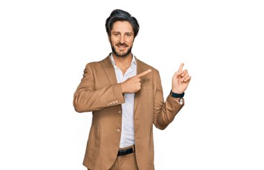 İş kıyafetleri giymiş, gülümseyen ve kameraya bakan genç İspanyol adam iki eliyle ve parmaklarıyla kenarı işaret ediyor.. 