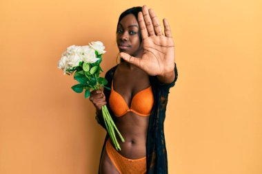 Genç Afrikalı Amerikalı kadın elinde çiçekle elinde iç çamaşırlarıyla dur işareti yapıyor ciddi ve kendinden emin bir ifadeyle, savunma jesti yapıyor. 