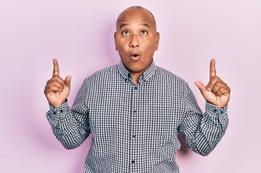 Orta yaşlı, gündelik kıyafetler giyen Latin bir adam şaşırmış ve şaşırmış bir şekilde yukarı bakıyor ve parmaklarıyla işaret ediyor ve kollarını kaldırıyor.. 