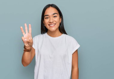 Genç Asyalı kadın günlük beyaz tişört giyer ve üç numaralı parmaklarıyla gülümseyerek mutlu ve kendinden emin bir şekilde işaret eder.. 