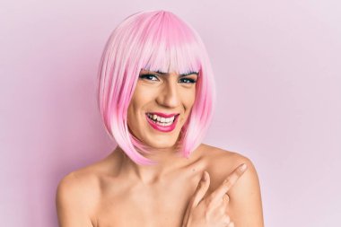 Pembe peruk takan genç adam mutlu ve doğal bir ifadeyle elleri ve parmaklarıyla gülümseyen neşeli bir yüzle makyaj yapıyor. 