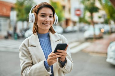Genç İspanyol kadın şehirde akıllı telefon ve kulaklık kullanarak mutlu bir şekilde gülümsüyor..