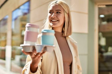 Genç sarışın kız gülümsüyor. Mutluluk tepsisi tutuyor. Şehirde kahve alıyor..