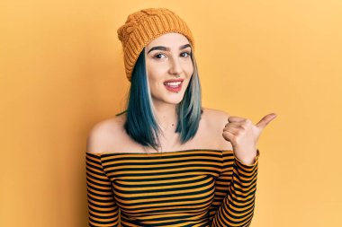 Yün şapka takan, gülümseyen, mutlu yüzlü genç bir kız başparmağıyla yana bakıyor ve işaret ediyor.. 