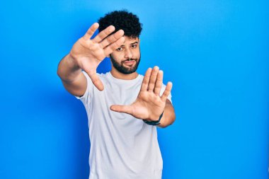 Sakallı, günlük beyaz tişörtlü, el ve parmaklarıyla çerçeve yapan genç bir Arap. 