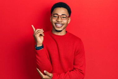 Genç Afro-Amerikan adam günlük kıyafetler ve gözlük takıyor, yüzünde kocaman bir gülümseme var, eli ve parmağı kameraya bakıyor.. 