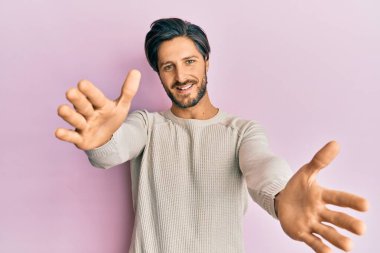 Latin asıllı genç adam, sıradan bir kazak giyip kameraya bakıyor ve sarılmak için kollarını açmış gülümsüyor. Mutluluğu kucaklayan neşeli bir ifade.. 