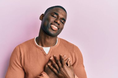 Sıradan kıyafetler giyen, göğsünde elleri kapalı, yüzünde minnet dolu jestlerle gülümseyen Afro-Amerikan bir genç. sağlık kavramı. 