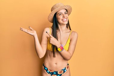 Bikini giyen ve yaz şapkası takan genç İspanyol kadın kameraya hayret ediyor ve gülümsüyor.. 