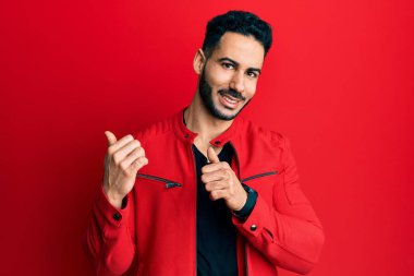 Kırmızı deri ceket giyen genç İspanyol adam, arka tarafı işaret ediyor. El ve baş parmakları havada, kendinden emin gülümsüyor. 