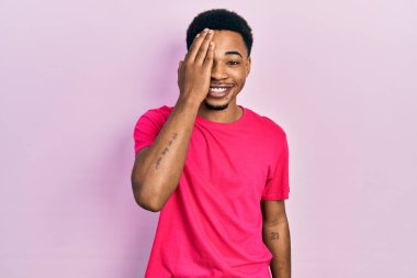 Genç Afrikalı Amerikalı adam Casua tişörtü giyiyor. Bir gözünü eliyle kapatıyor. Yüzünde kendine güvenen bir gülümseme ve şaşırtıcı bir duygu var.. 