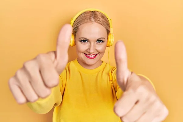 ヘッドフォンを使用して音楽を聴いている若いブロンドの女性は 手で積極的なジェスチャーを行うことを承認し 親指を上げて笑顔と成功のために幸せ 勝者のジェスチャー — ストック写真