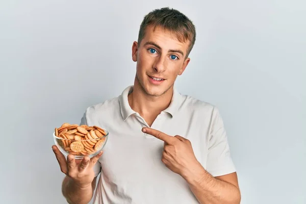 Jovem Caucasiano Segurando Tigela Biscoitos Salgados Sorrindo Feliz Apontando Com — Fotografia de Stock