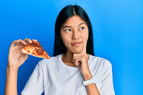 年轻的中国女人吃着美味的辣香肠披萨 严肃的脸上带着下巴思考着问题 思考着困惑的想法 — 图库照片