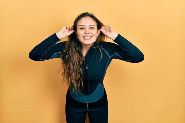 Young hispanic girl wearing diver neoprene uniform smiling pulling ears with fingers, funny gesture. audition problem