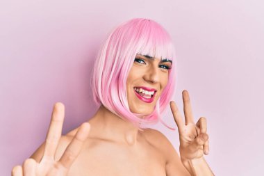 Pembe peruk takan, gülümseyen, parmaklarını zafer işareti yaparken gösteren genç bir adam. İki numara.. 