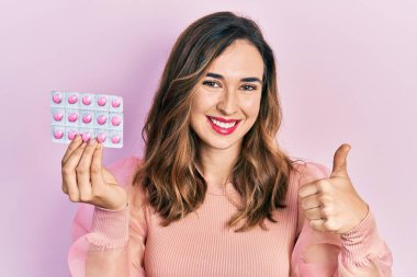 Genç İspanyol kız elinde haplarla mutlu ve pozitif gülümsüyor, başparmağıyla mükemmel ve onay işareti yapıyor. 