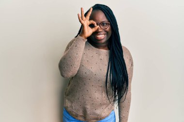 Saçları örgülü, günlük kıyafetler ve gözlük takan genç siyahi bir kadın el hareketleriyle gülümsüyor ve mutlu yüzüyle parmaklarının arasından bakıyor.. 