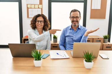 Orta yaşlı İspanyol kadın ve erkek ofiste dizüstü bilgisayarla oturmuş kameraya hayret ediyor ve gülümsüyor.. 