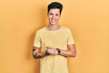 Sarı günlük tişörtler giyen, ellerini kavuşturup gülümseyen, gevşemiş ve neşeli genç İspanyol bir adam. Başarı ve iyimserlik 