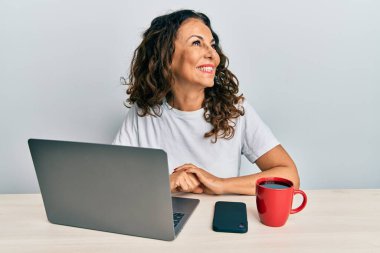 Ofiste çalışan orta yaşlı güzel bir kadın bilgisayarını kullanıyor. Yüzünde gülümseme ve doğal ifadeyle yan yana bakıyor. Kendinden emin gülen. 