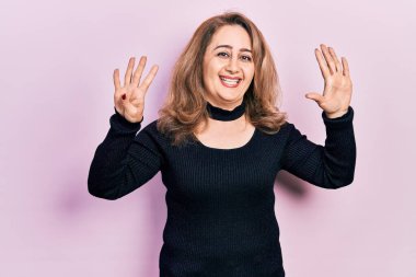 Orta yaşlı beyaz kadın, günlük kıyafetler giyip dokuz numaralı parmaklarıyla işaret ederken kendinden emin ve mutlu bir şekilde gülümsüyor.. 