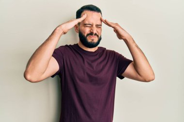 Sakallı genç bir adam, elleri kafasında rahat bir tişört giyiyor, stresten baş ağrısı çekiyor. migren ağrısı. 