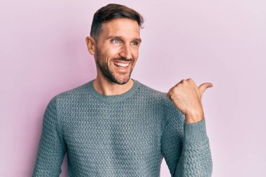 Kış süveteri giymiş sakallı yakışıklı bir adam başparmağını yana uzatıyor ve ağzı açık gülümsüyor. 