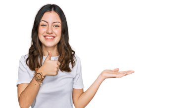 Gayri resmi beyaz tişört giyen genç ve güzel bir kadın avuç içini gösteriyor ve baş parmaklarıyla iyi bir jest yapıyor, mutlu ve neşeli gülümsüyor. 