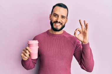 Sakallı genç adam bir fincan kahve içiyor tamam işareti yapıyor parmaklarıyla, gülümseyerek mükemmel bir sembol oluşturuyor. 