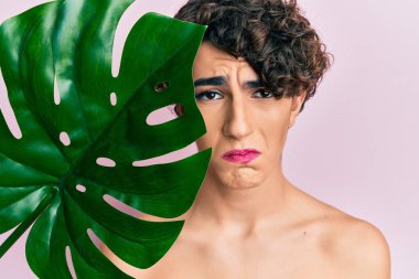 Güzel yüzüne yakın yeşil yaprak tutan, makyaj yapan genç bir adam depresif ve endişeli, sinirli ve korkmuş bir halde. hüzünlü ifade. 