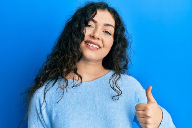 Kıvırcık saçlı, günlük kıyafetler giyen genç esmer kadın mutlu bir el hareketi yapıyor. Kameraya bakıp başarıyı gösteren ifade. 