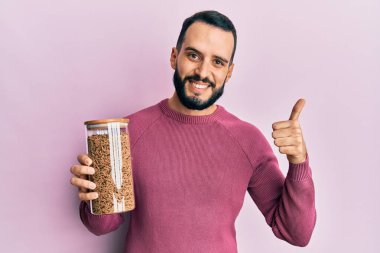 Sakallı genç adam sağlıklı tahıl gevreği kavanozunu tutuyor mutlu ve pozitif gülümsüyor, başparmağı mükemmel ve onay işareti yapıyor. 