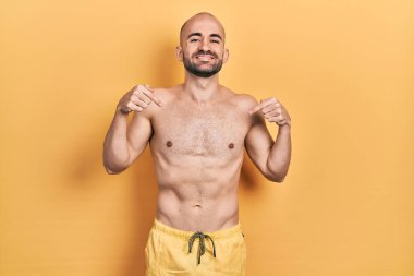 Yüzünde bir gülümsemeyle mayo giyen genç ve kel bir adam kendini gururlu ve mutlu olarak gösteriyor.. 