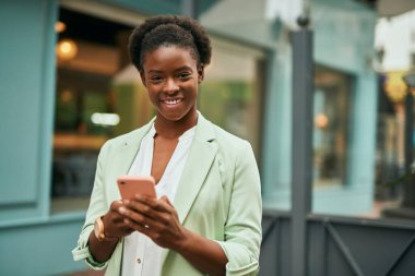 Genç Afrikalı Amerikalı iş kadını şehirde akıllı telefon kullanarak mutlu bir şekilde gülümsüyor..