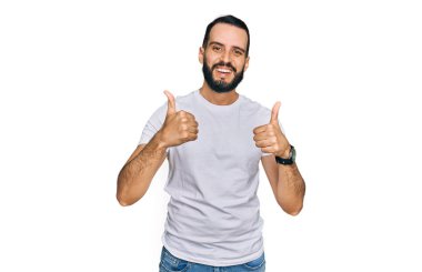 Sakallı genç adam, günlük beyaz tişört giyiyor. Başarı işareti yapıyor. Elle olumlu jest yapıyor. Neşeli ifade ve kazanan jest. 