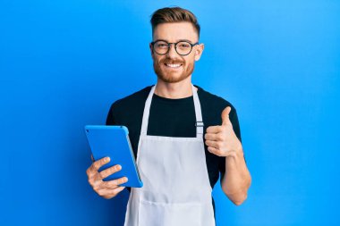 Genç kızıl saçlı adam profesyonel önlük giyiyor dokunmatik ped kullanıyor mutlu ve pozitif gülümsüyor, başparmağıyla mükemmel ve onay işareti yapıyor. 