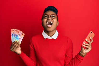 Genç Afrikalı Amerikalı kadın elinde İsviçre frangı banknotları ile akıllı telefon kullanıyor kızgın ve kızgın çığlıklar atıyor öfkeyle bağırıyor, öfkeyle yukarı bakıyor. 
