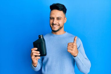Motor yağı şişesi tutan genç Arap adam mutlu ve pozitif gülümsüyor, başparmağıyla mükemmel ve onay işareti yapıyor. 