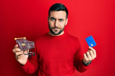 Genç İspanyol adam elinde küçük bir süpermarket alışveriş arabası ve kredi kartı şüphecisi ve gergin, sorun yüzünden somurtuyor. negatif kişi. 