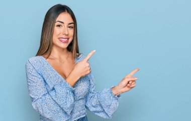 Gündelik kıyafetler giyen, gülümseyen ve kameraya bakan genç İspanyol kadın iki eliyle ve parmaklarıyla kenarı işaret ediyor.. 