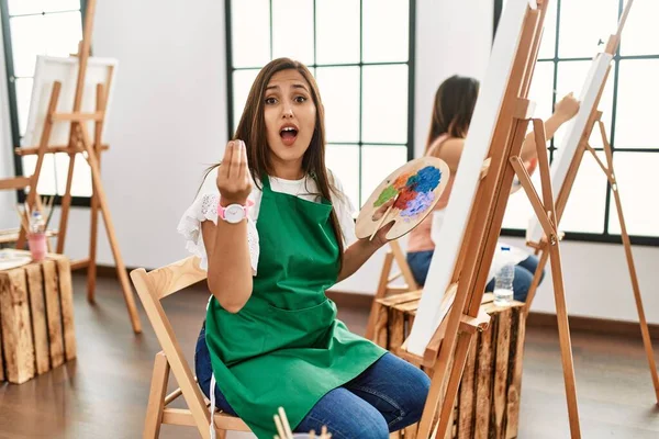 若いヒスパニックアーティストの女性は 手と指で自信を持って表現してイタリアのジェスチャーを行うアートスタジオでキャンバスに絵を描く — ストック写真