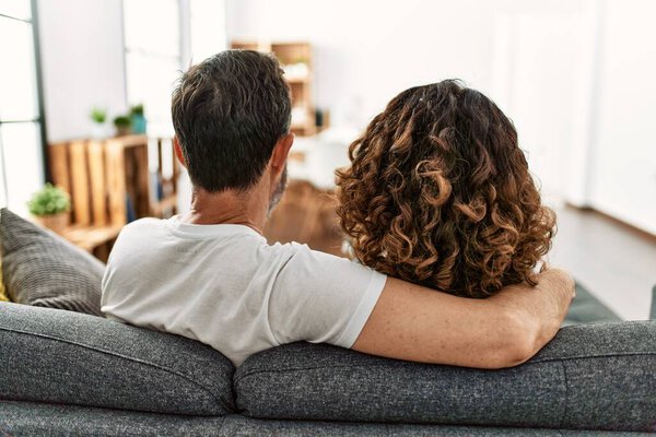 Middle age hispanic couple on back view hugging and sitting on the sofa at home.