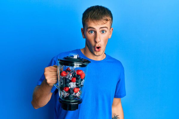 Young caucasian man making smoothie holding electric mixer scared and amazed with open mouth for surprise, disbelief face 
