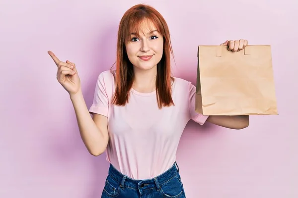 Pelirroja Mujer Joven Sosteniendo Llevar Bolsa Papel Sonriendo Feliz Señalando — Foto de Stock