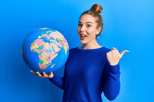 Young Blonde Woman Holding World Ball Pointing Thumb Side Smiling — Stock Photo, Image